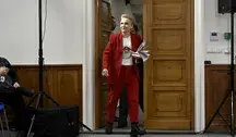 Joanna Scheuring-Wielgus wchodzi do sali konferencyjnej w czerwonym garniturze, trzymając dokumenty. Zdjęcie ilustruje jej udział w debacie publicznej w kontekście krytyki decyzji prezydenta Karola Nawrockiego o odebraniu Orderu Orła Białego Wołodymyrowi Zełenskiemu