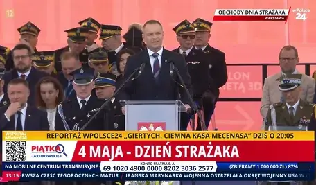 Prezydent Karol Nawrocki przemawia podczas obchodów Dnia Strażaka na placu w Warszawie. W tle widoczna grupa strażaków, w tym funkcjonariusze Państwowej Straży Pożarnej oraz wysokiej rangi wojskowi, z różnorodnymi odznaczeniami i mundurami. Na pierwszym planie widoczna jest scena z mikrofonem, a nad nią napis „Obchody Dnia Strażaka”. W tle czerwony baner z symbolem Polski