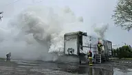 Fotografia przedstawia akcję gaśniczą strażaków, którzy walczą z pożarem kontenera z magazynem energii. Gęsty dym wznosi się ku niebu, a strażacy w pełnym ekwipunku starają się opanować płomienie za pomocą wody i specjalistycznego sprzętu. Pożar wybuchł w wyniku awarii akumulatorów litowo-jonowych, które przechowywały energię odnawialną. W działaniach bierze udział 27 zastępów straży pożarnej.