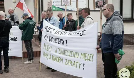 Grupa protestujących pracowników spółki FIBRIS trzymających transparenty z hasłami o dramatycznej sytuacji związanej z brakiem wypłat i blokadą kont przez ZUS. Na pierwszym planie widoczna jest biało-zielona flaga Polski oraz transparent z napisem: „PRZEZ WASZE DECYZJE MY GŁODUJEMY! ŻYJEMY W ZAWIESZENIU MIĘDZY PRACĄ A BRAKIEM ZAPŁATY!” Protest odbywa się przed siedzibą ZUS w Rzeszowie