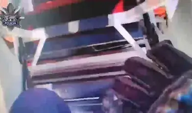 Klatka z nagrania policyjnego przedstawiająca wnętrze radiowozu podczas interwencji, widoczne elementy wyposażenia pojazdu oraz dynamiczna scena z akcji służb Policji.