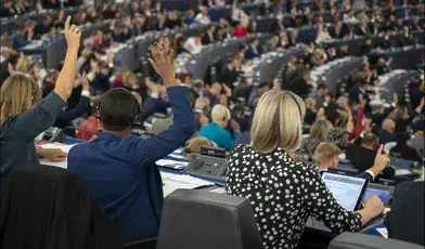 Sala obrad Parlamentu Europejskiego podczas głosowania, widok od tyłu na delegatów siedzących przy stanowiskach z mikrofonami i komputerami. Część europosłów podnosi ręce w głosowaniu, w tle widoczne liczne rzędy uczestników sesji plenarnej.
