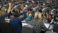 Sala obrad Parlamentu Europejskiego podczas głosowania, widok od tyłu na delegatów siedzących przy stanowiskach z mikrofonami i komputerami. Część europosłów podnosi ręce w głosowaniu, w tle widoczne liczne rzędy uczestników sesji plenarnej.