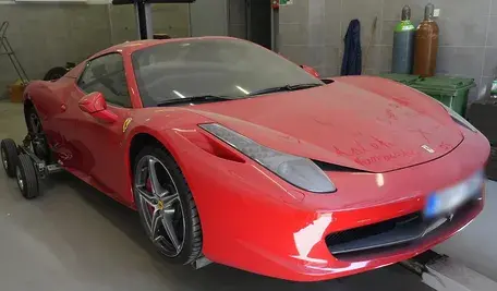 Czerwone Ferrari zabezpieczone przez policję, ustawione w warsztacie na wózkach transportowych, z zamazanymi elementami nadwozia i widocznymi napisami na masce.