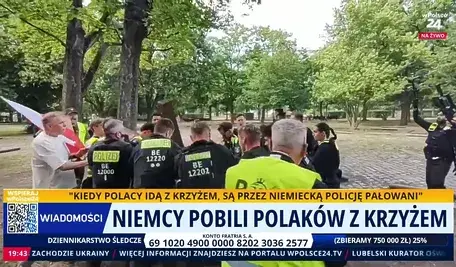 Funkcjonariusze niemieckiej policji w kamizelkach odblaskowych otaczają grupę osób w parku miejskim. Wśród uczestników widoczna flaga Polski oraz drewniany krzyż, część osób rozmawia z policją podczas interwencji.