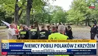 Funkcjonariusze niemieckiej policji w kamizelkach odblaskowych otaczają grupę osób w parku miejskim. Wśród uczestników widoczna flaga Polski oraz drewniany krzyż, część osób rozmawia z policją podczas interwencji.