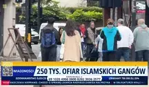 Ujęcie z ulicy przedstawiające grupę przechodniów na chodniku w mieście. Na dole ekranu widoczny pasek informacyjny programu telewizyjnego z napisem „250 tys. ofiar islamskich gangów” oraz elementami graficznymi stacji informacyjnej.