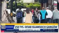 Ujęcie z ulicy przedstawiające grupę przechodniów na chodniku w mieście. Na dole ekranu widoczny pasek informacyjny programu telewizyjnego z napisem „250 tys. ofiar islamskich gangów” oraz elementami graficznymi stacji informacyjnej.