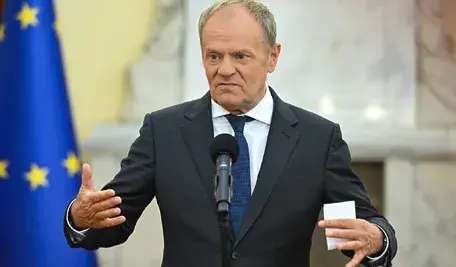 Donald Tusk przemawiający podczas konferencji prasowej przy mównicy z mikrofonem, wykonujący gesty rękami. W tle widoczna flaga Unii Europejskiej, scena w budynku instytucji publicznej.