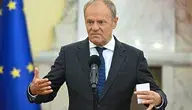 Donald Tusk przemawiający podczas konferencji prasowej przy mównicy z mikrofonem, wykonujący gesty rękami. W tle widoczna flaga Unii Europejskiej, scena w budynku instytucji publicznej.