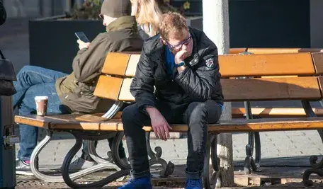 Zrezygnowany młody mężczyzna siedzi na ławce w mieście, z głową opartą na dłoni, ilustrując problem bezrobocia i rosnących długów wśród młodych Polaków