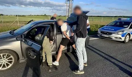 Policjanci zatrzymują podejrzanego mężczyznę przy samochodzie osobowym na drodze. W tle widoczny jest radiowóz oraz drugi zatrzymany.