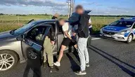 Policjanci zatrzymują podejrzanego mężczyznę przy samochodzie osobowym na drodze. W tle widoczny jest radiowóz oraz drugi zatrzymany.