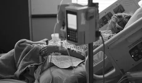 Pacjentka leżąca w szpitalnym łóżku podłączona do aparatury medycznej, w tym kroplówki i monitorów funkcji życiowych. Ujęcie wykonane w czerni i bieli podkreśla powagę sytuacji oraz atmosferę opieki medycznej i hospitalizacji.