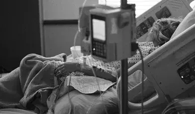 Pacjentka leżąca w szpitalnym łóżku podłączona do aparatury medycznej, w tym kroplówki i monitorów funkcji życiowych. Ujęcie wykonane w czerni i bieli podkreśla powagę sytuacji oraz atmosferę opieki medycznej i hospitalizacji.