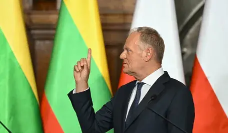 Donald Tusk podczas wystąpienia publicznego na konferencji, gestykulujący uniesionym palcem w trakcie przemówienia. W tle widoczne są flagi Polski i Litwy, sugerujące kontekst spotkania międzynarodowego lub dwustronnych rozmów.
