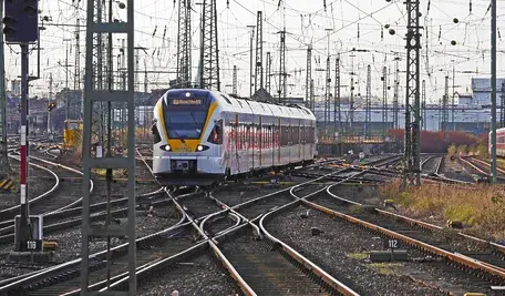 Nowoczesny pociąg pasażerski wjeżdżający na rozbudowany węzeł kolejowy w Niemczech, otoczony gęstą siecią torów, rozjazdów i trakcji elektrycznej. W tle widoczne są słupy sieci trakcyjnej, sygnalizacja oraz inne składy kolejowe stojące na bocznych torach. Scena przedstawia złożoną infrastrukturę kolejową i ruch pociągów na dużej stacji rozrządowej.