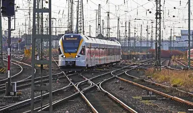 Nowoczesny pociąg pasażerski wjeżdżający na rozbudowany węzeł kolejowy w Niemczech, otoczony gęstą siecią torów, rozjazdów i trakcji elektrycznej. W tle widoczne są słupy sieci trakcyjnej, sygnalizacja oraz inne składy kolejowe stojące na bocznych torach. Scena przedstawia złożoną infrastrukturę kolejową i ruch pociągów na dużej stacji rozrządowej.
