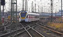 Nowoczesny pociąg pasażerski wjeżdżający na rozbudowany węzeł kolejowy w Niemczech, otoczony gęstą siecią torów, rozjazdów i trakcji elektrycznej. W tle widoczne są słupy sieci trakcyjnej, sygnalizacja oraz inne składy kolejowe stojące na bocznych torach. Scena przedstawia złożoną infrastrukturę kolejową i ruch pociągów na dużej stacji rozrządowej.