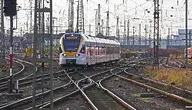 Nowoczesny pociąg pasażerski wjeżdżający na rozbudowany węzeł kolejowy w Niemczech, otoczony gęstą siecią torów, rozjazdów i trakcji elektrycznej. W tle widoczne są słupy sieci trakcyjnej, sygnalizacja oraz inne składy kolejowe stojące na bocznych torach. Scena przedstawia złożoną infrastrukturę kolejową i ruch pociągów na dużej stacji rozrządowej.