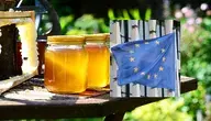 Słoiki z miodem i plaster miodu na stole ogrodowym, obok flaga Unii Europejskiej, symbolizujące nowe unijne przepisy dotyczące etykietowania i jakości miodu