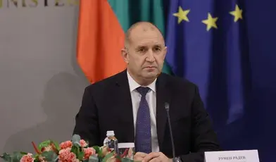 Premier Bułgarii Rumen Radev podczas konferencji prasowej, siedzi przy stole z mikrofonem i kwiatami, w tle flagi Bułgarii i UE.