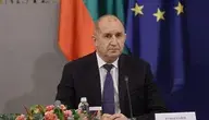 Premier Bułgarii Rumen Radev podczas konferencji prasowej, siedzi przy stole z mikrofonem i kwiatami, w tle flagi Bułgarii i UE.