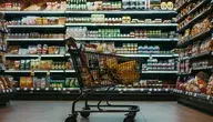 Wózek sklepowy w supermarkecie na tle półek pełnych produktów spożywczych, symbolizujący zakupy żywności w sklepie.
