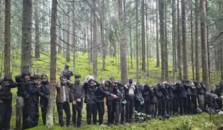 Grupa zatrzymanych cudzoziemców w lesie w południowo-wschodniej Łotwie, w pobliżu granicy z Białorusią. Mężczyźni i kobiety w cywilnych ubraniach, wśród nich strażnicy graniczni w mundurach. W tle widać gęste zarośla i drzewa, w ziemi zamaskowane wejście do podziemnego tunelu, którym przedostali się z Białorusi. Akcja miała miejsce 20 sierpnia 2026 roku w gminie Krasław