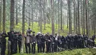 Grupa zatrzymanych cudzoziemców w lesie w południowo-wschodniej Łotwie, w pobliżu granicy z Białorusią. Mężczyźni i kobiety w cywilnych ubraniach, wśród nich strażnicy graniczni w mundurach. W tle widać gęste zarośla i drzewa, w ziemi zamaskowane wejście do podziemnego tunelu, którym przedostali się z Białorusi. Akcja miała miejsce 20 sierpnia 2026 roku w gminie Krasław