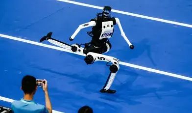 Humanoidalny robot biegnie po niebieskiej bieżni podczas World Humanoid Robot Games na stadionie Ice Ribbon w Pekinie. W tle widoczni są operatorzy maszyn i publiczność obserwująca zawody