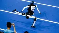 Humanoidalny robot biegnie po niebieskiej bieżni podczas World Humanoid Robot Games na stadionie Ice Ribbon w Pekinie. W tle widoczni są operatorzy maszyn i publiczność obserwująca zawody
