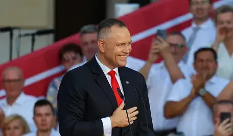 Prezydent RP Karol Nawrocki podczas wystąpienia na dziedzińcu Pałacu Prezydenckiego w pierwszą rocznicę zaprzysiężenia. W tle scena, publiczność oraz biało-czerwone flagi.