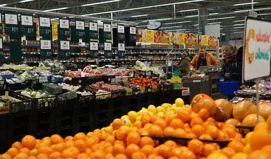 Sklepowe stoisko z produktami spożywczymi – na półkach pieczywo, nabiał, słodycze. Wyraźnie widoczne metki z cenami, niektóre z przekreślonymi starymi kwotami i nowymi, wyższymi. Ilustracja codziennej rzeczywistości zakupów Polaków w czasach inflacji.