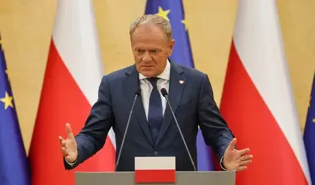 Premier Donald Tusk w ciemnym garniturze i krawacie gestykuluje obiema rękami za mównicą na tle flag Polski i Unii Europejskiej.