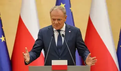 Premier Donald Tusk w ciemnym garniturze i krawacie gestykuluje obiema rękami za mównicą na tle flag Polski i Unii Europejskiej.
