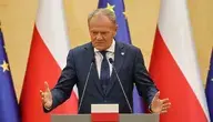 Premier Donald Tusk w ciemnym garniturze i krawacie gestykuluje obiema rękami za mównicą na tle flag Polski i Unii Europejskiej.
