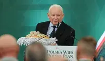 Jarosław Kaczyński w ciemnym garniturze stoi za białą mównicą z napisem „Polska Wieś Polski Plan”. Przed nim leży tradycyjny, ozdobny bochenek chleba na serwecie. W tle widoczna zielona ścianka z grafiką.