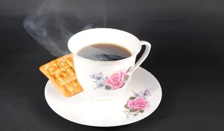Biała porcelanowa filiżanka z wzorem róż, wypełniona gorącą, parującą czarną kawą. Obok filiżanki na talerzyku leży jeden prostokątny herbatnik. Całość na ciemnym, kontrastowym tle.
