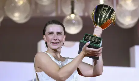 Wzruszona duńska aktorka Mathilde Arcel w białej sukience, z uśmiechem trzymająca w obu dłoniach wzniesiony złoty puchar Volpiego dla najlepszej aktorki na scenie Festiwalu Filmowego w Wenecji. W tle widać rozmyte, ozdobne kuliste lampy.