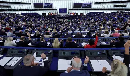 Szeroki kadr na salę plenarną Parlamentu Europejskiego w Strasburgu. Setki eurodeputowanych siedzących w półokrągłych rzędach przy niebieskich pulpitach. Część z nich ma podniesione ręce w trakcie trwającego głosowania. W centrum sali, za mównicą, widoczne są flagi państw członkowskich oraz flaga Unii Europejskiej.