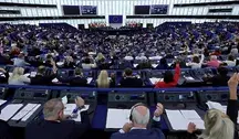 Szeroki kadr na salę plenarną Parlamentu Europejskiego w Strasburgu. Setki eurodeputowanych siedzących w półokrągłych rzędach przy niebieskich pulpitach. Część z nich ma podniesione ręce w trakcie trwającego głosowania. W centrum sali, za mównicą, widoczne są flagi państw członkowskich oraz flaga Unii Europejskiej.