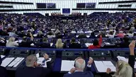 Szeroki kadr na salę plenarną Parlamentu Europejskiego w Strasburgu. Setki eurodeputowanych siedzących w półokrągłych rzędach przy niebieskich pulpitach. Część z nich ma podniesione ręce w trakcie trwającego głosowania. W centrum sali, za mównicą, widoczne są flagi państw członkowskich oraz flaga Unii Europejskiej.
