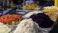 Stołówkowy bufet z warzywami i dodatkami do posiłków, m.in. pomidorami, ogórkami, surówką z czerwonej kapusty, kukurydzą i serem feta