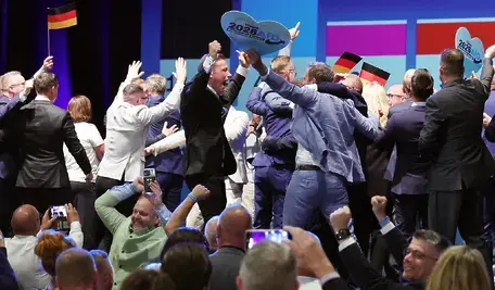 Uczestnicy wydarzenia AfD świętują na scenie z niemieckimi flagami i tablicami „Vision 2026 – Deutschland zuerst”.