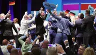 Uczestnicy wydarzenia AfD świętują na scenie z niemieckimi flagami i tablicami „Vision 2026 – Deutschland zuerst”.