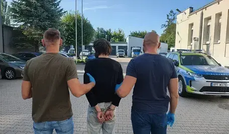 Dwóch mężczyzn w cywilnych ubraniach prowadzi zatrzymanego, zakutego w kajdanki z tyłu, mężczyznę w ciemnej bluzie i dżinsach. Tło stanowi parking policyjny z radiowozami.