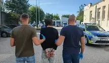 Dwóch mężczyzn w cywilnych ubraniach prowadzi zatrzymanego, zakutego w kajdanki z tyłu, mężczyznę w ciemnej bluzie i dżinsach. Tło stanowi parking policyjny z radiowozami.