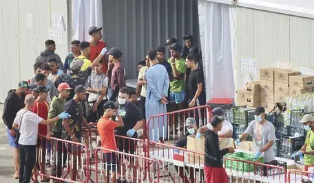Grupa młodych migrantów w obozie tymczasowym w Ceucie przy barierkach odgradzających, obsługa w rękawiczkach i maseczkach wydaje zaopatrzenie