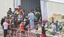 Grupa młodych migrantów w obozie tymczasowym w Ceucie przy barierkach odgradzających, obsługa w rękawiczkach i maseczkach wydaje zaopatrzenie